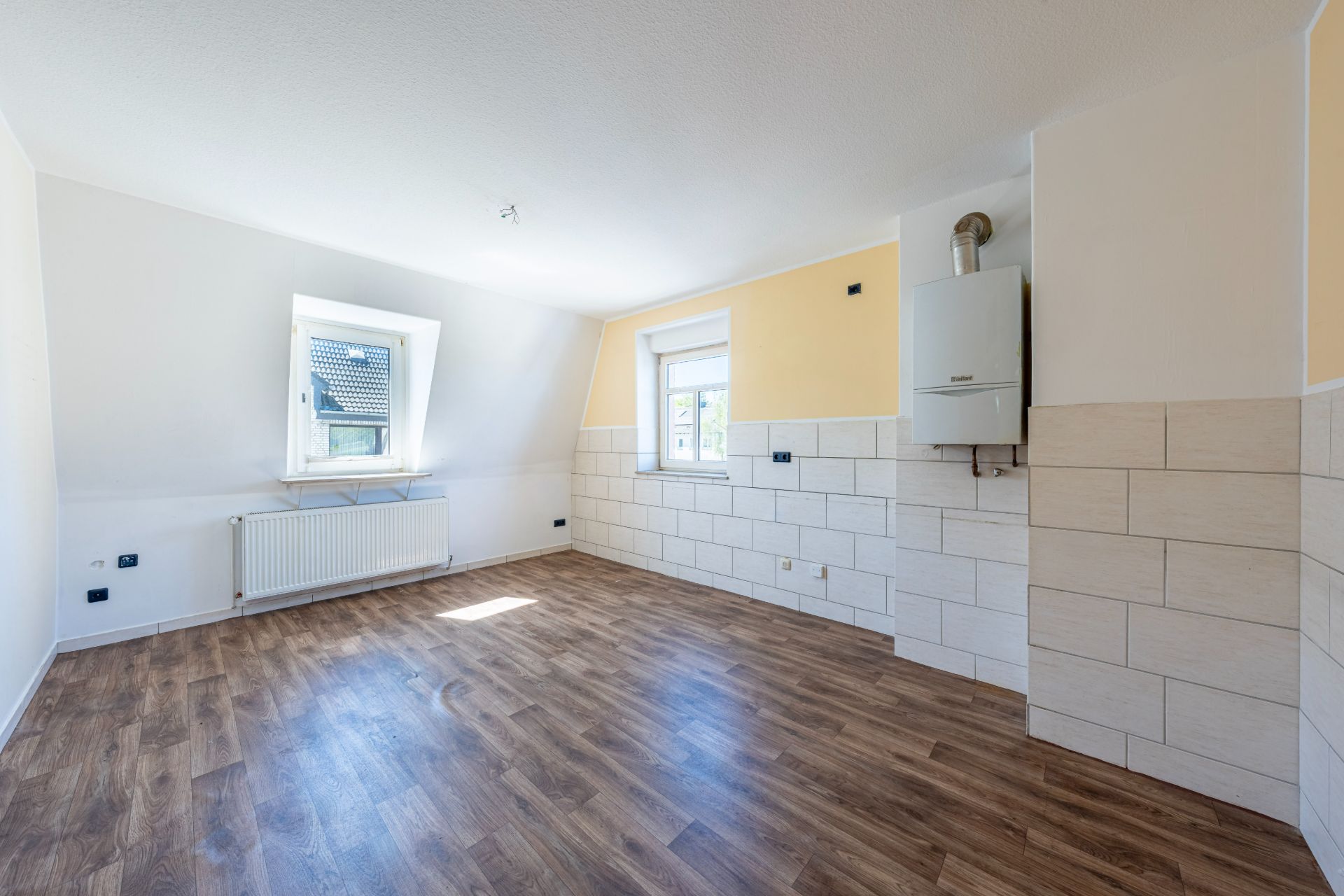 Heller leerer Raum mit zwei Fenstern, Holzboden und einer Heizanlage an der Wand.