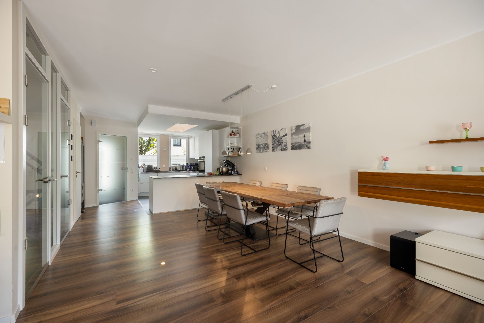 Helles Esszimmer mit großem Holztisch, sechs Stühlen, Holzboden und offener Küche im Hintergrund.