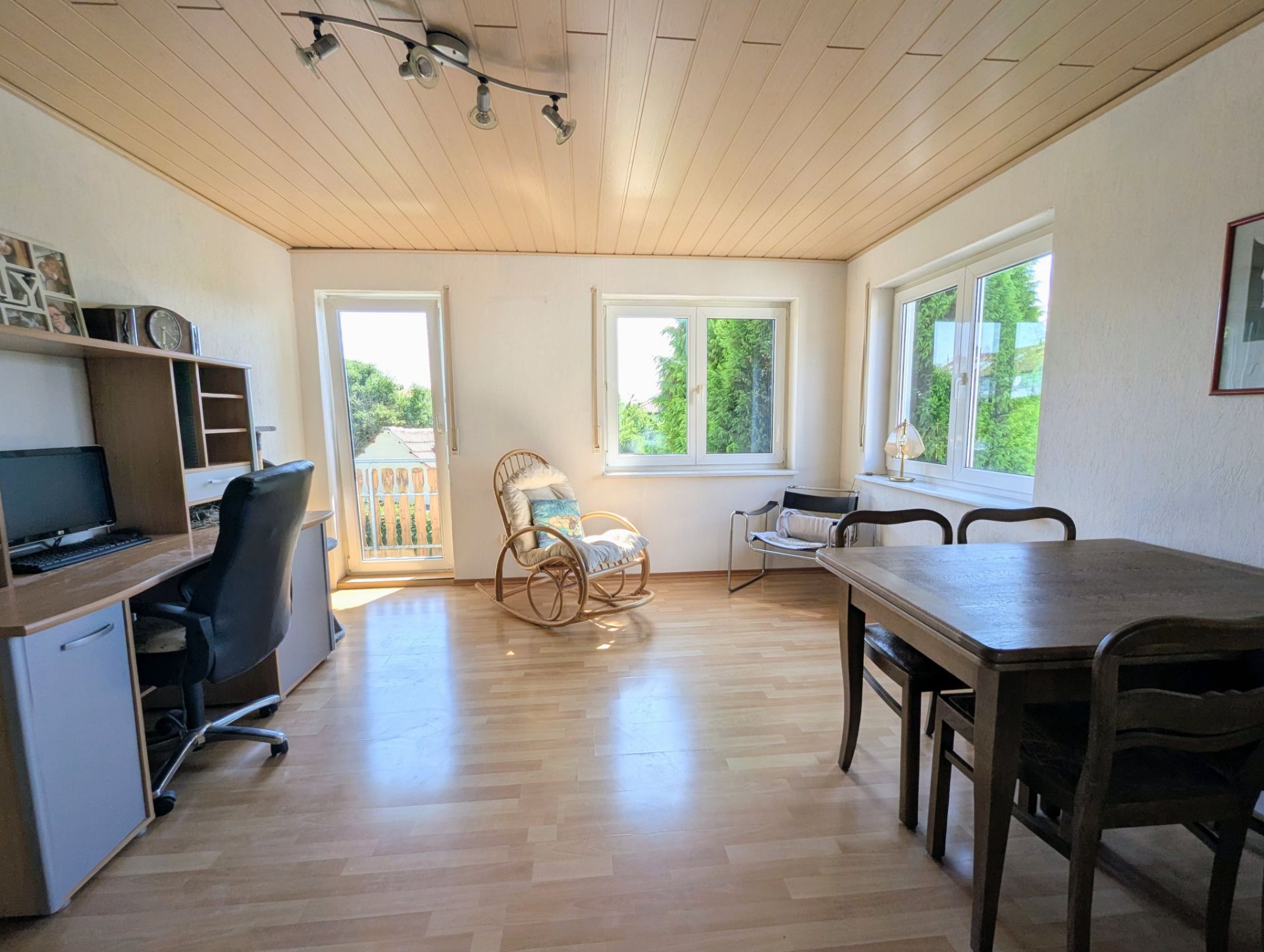 Helles Zimmer mit Schreibtisch, Stuhl, Esstisch, Korbsessel und großen Fenstern.