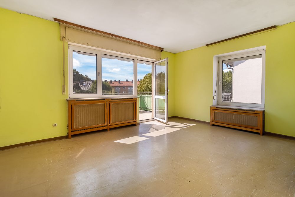 Leeres Zimmer mit gelben Wänden, zwei Fenstern und einer offenen Tür zum Balkon.