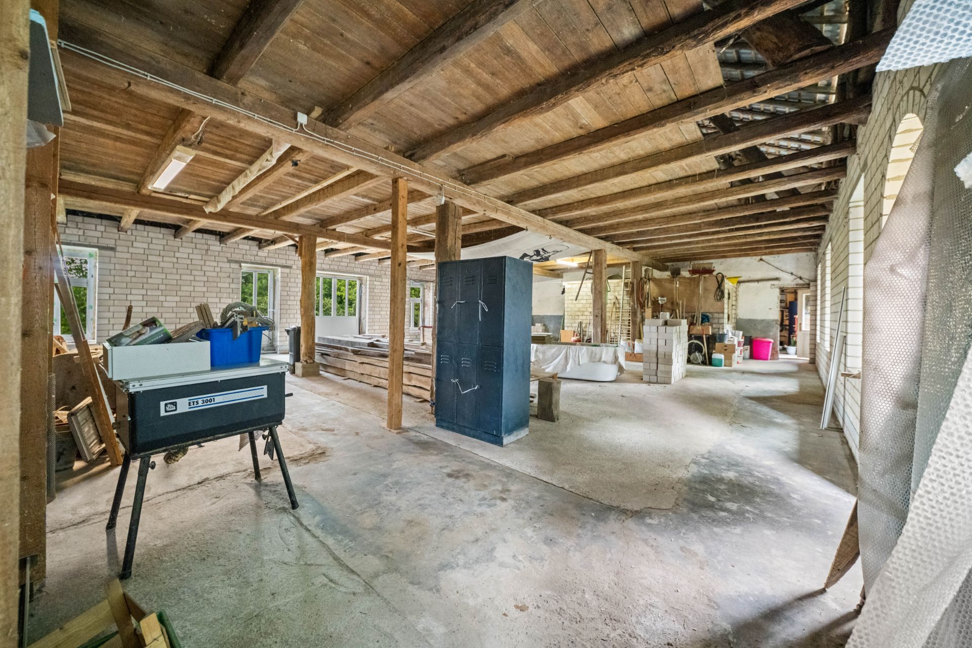 Großer, offener Raum mit sichtbaren Holzbalken, Werkstattgegenständen und Fenstern an den Wänden.