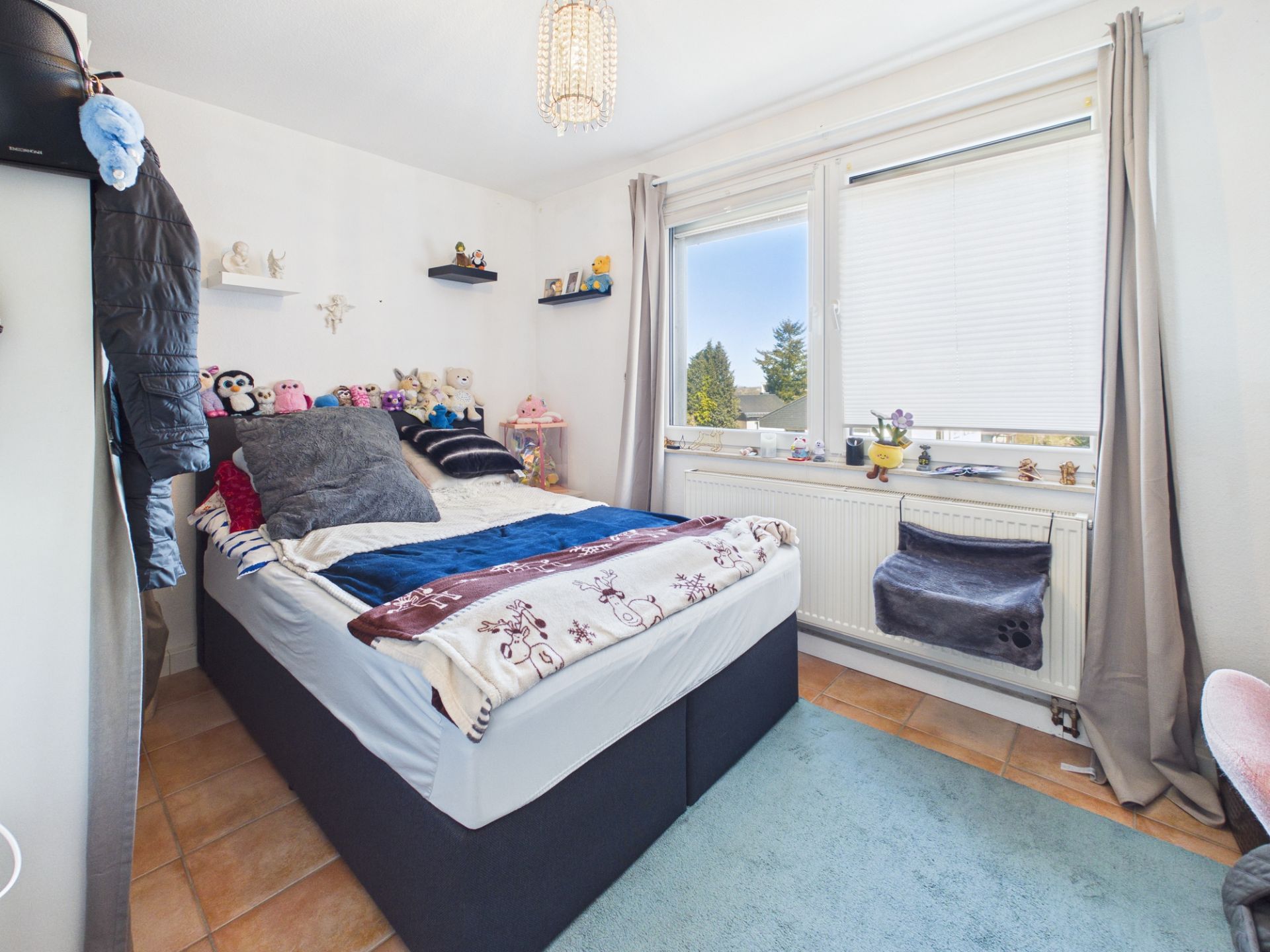 Kleines Schlafzimmer mit Bett, Kuscheltieren, Fenstern und Gardinen, Regal an der Wand.