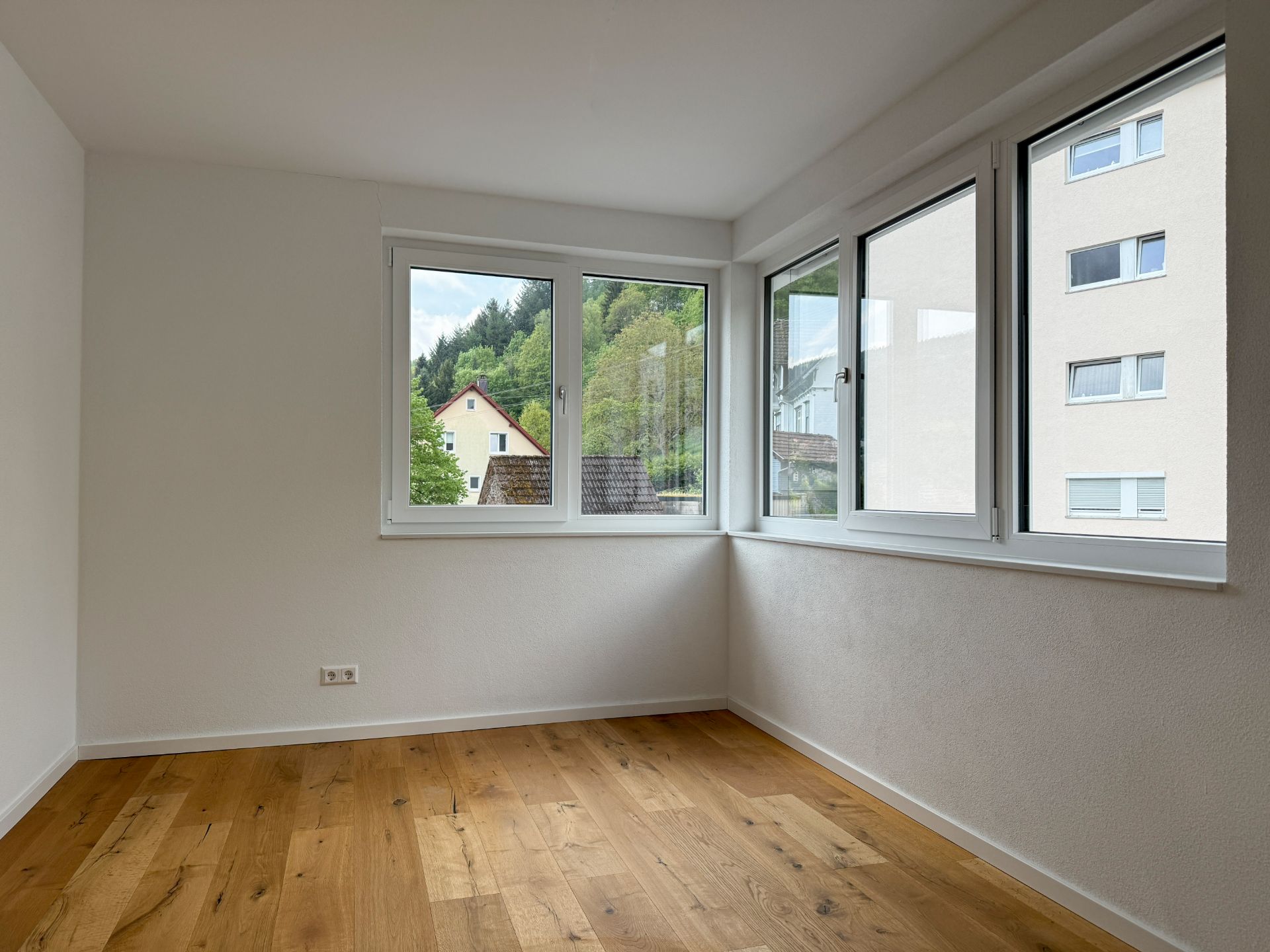 Leeres Zimmer mit Holzfußboden und großen Fenstern, Blick nach draußen auf Häuser und Bäume.