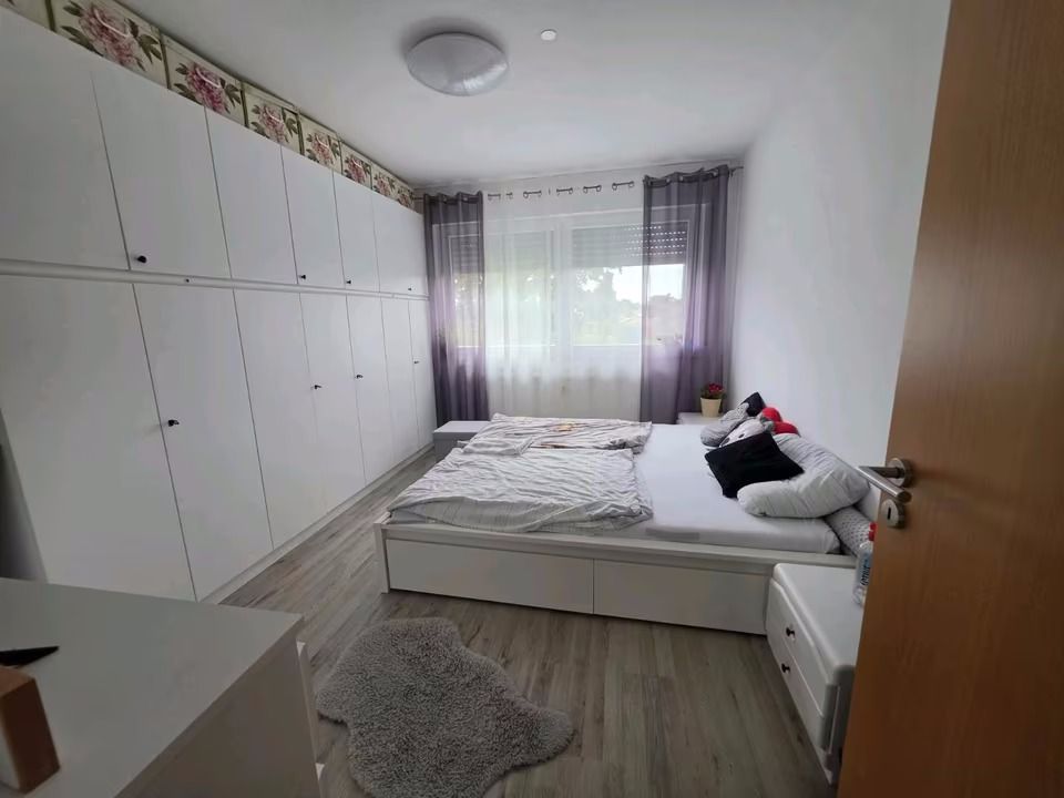 Helles Schlafzimmer mit großem weißen Kleiderschrank, Doppelbett und Fenster mit Gardinen.