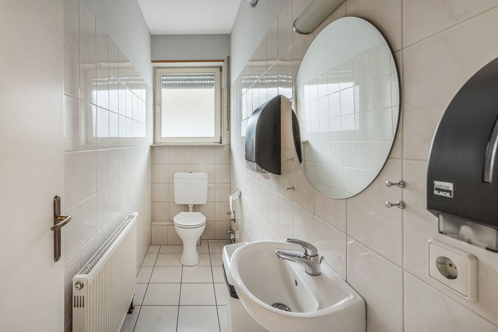 Schmales Badezimmer mit Toilette, kleinem Waschbecken, Spiegel und Fenster.