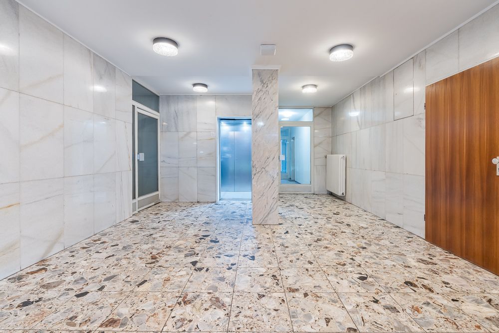 Heller Hausflur mit Marmorboden, weißen Wänden, Aufzug und Holztür.