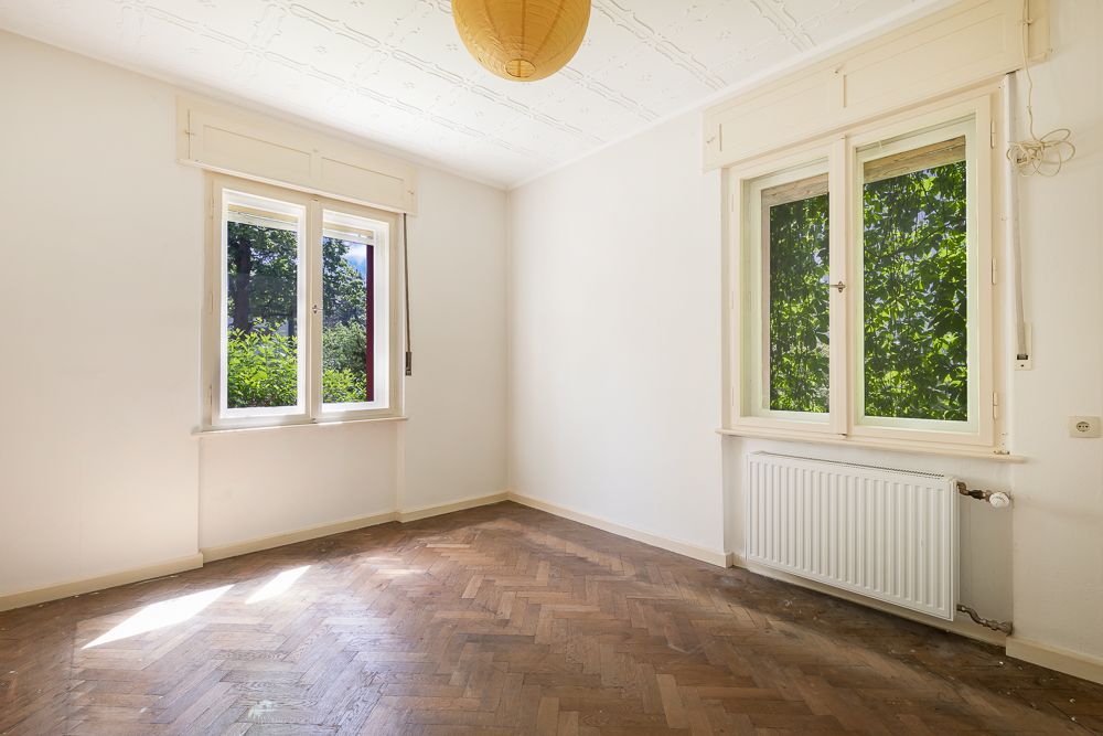 Leeres Zimmer mit Parkettboden, zwei Fenstern und Heizkörper an der Wand.