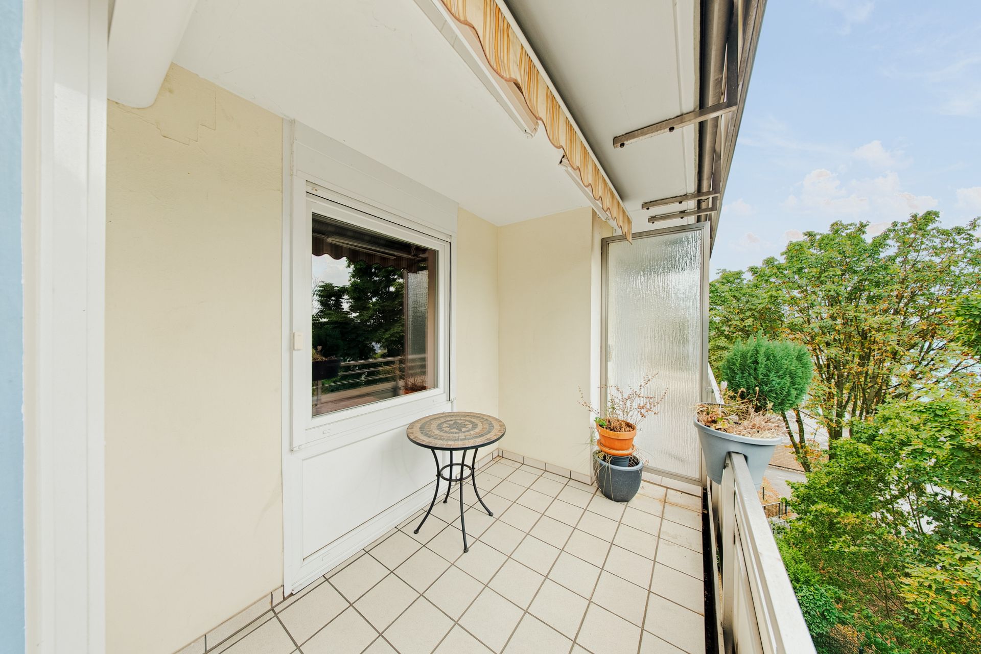 Überdachter Balkon mit Fliesenboden, kleinem Tisch, Pflanzen und Blick auf Bäume.