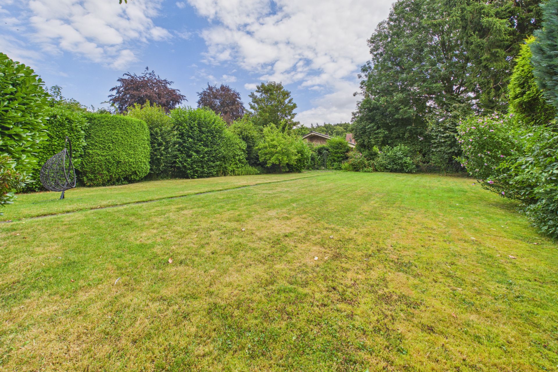 Großer, gepflegter Garten mit Rasenfläche, umgeben von Bäumen und Sträuchern, unter blauem Himmel.
