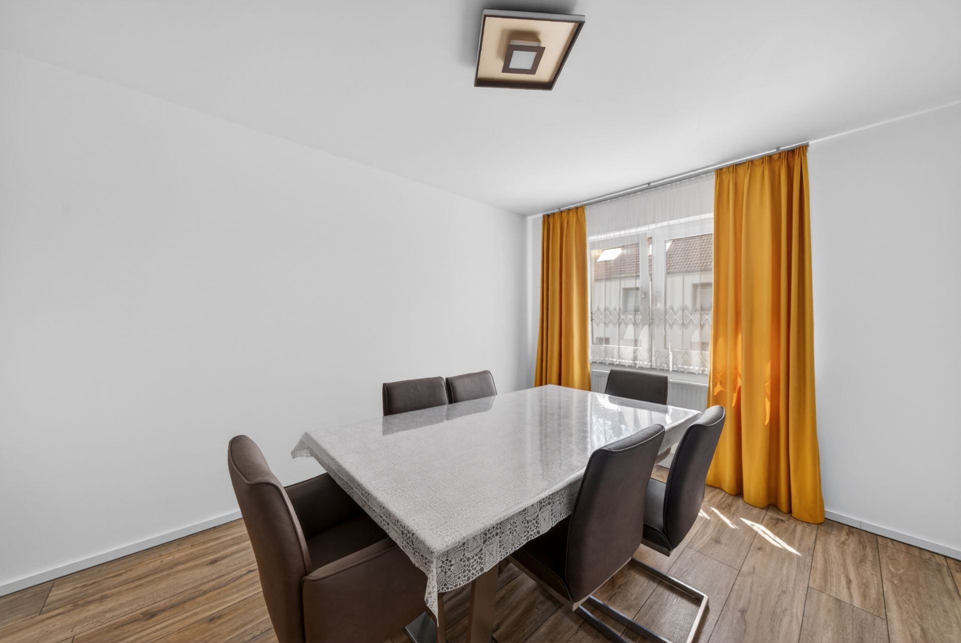 Esszimmer mit sechs Stühlen, Tischdecke und gelben Vorhängen vor dem Fenster.