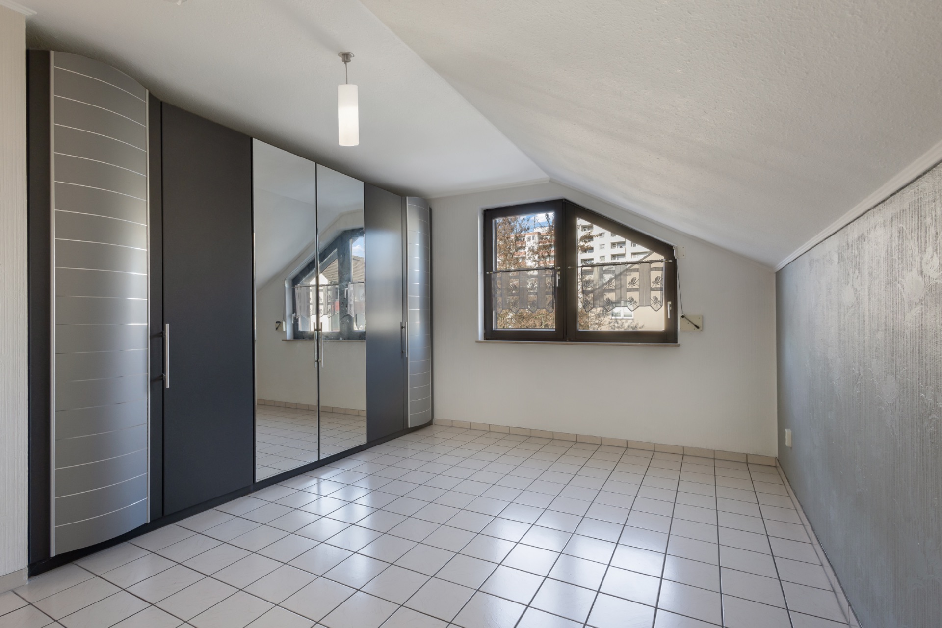 Leeres Zimmer mit Fliesenboden, großem Kleiderschrank und schrägem Fenster.