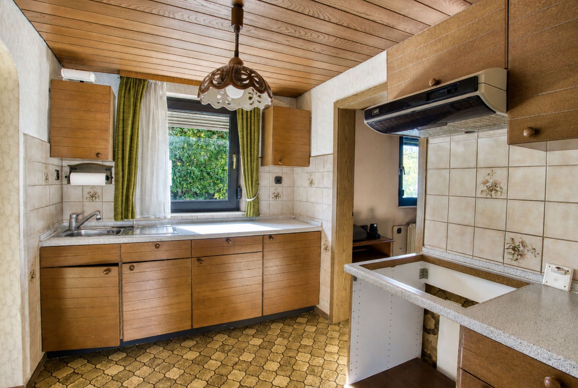 Küche mit Holzmöbeln, Fliesen, Spüle, Fenster mit grünen Vorhängen und Deckenlampe.