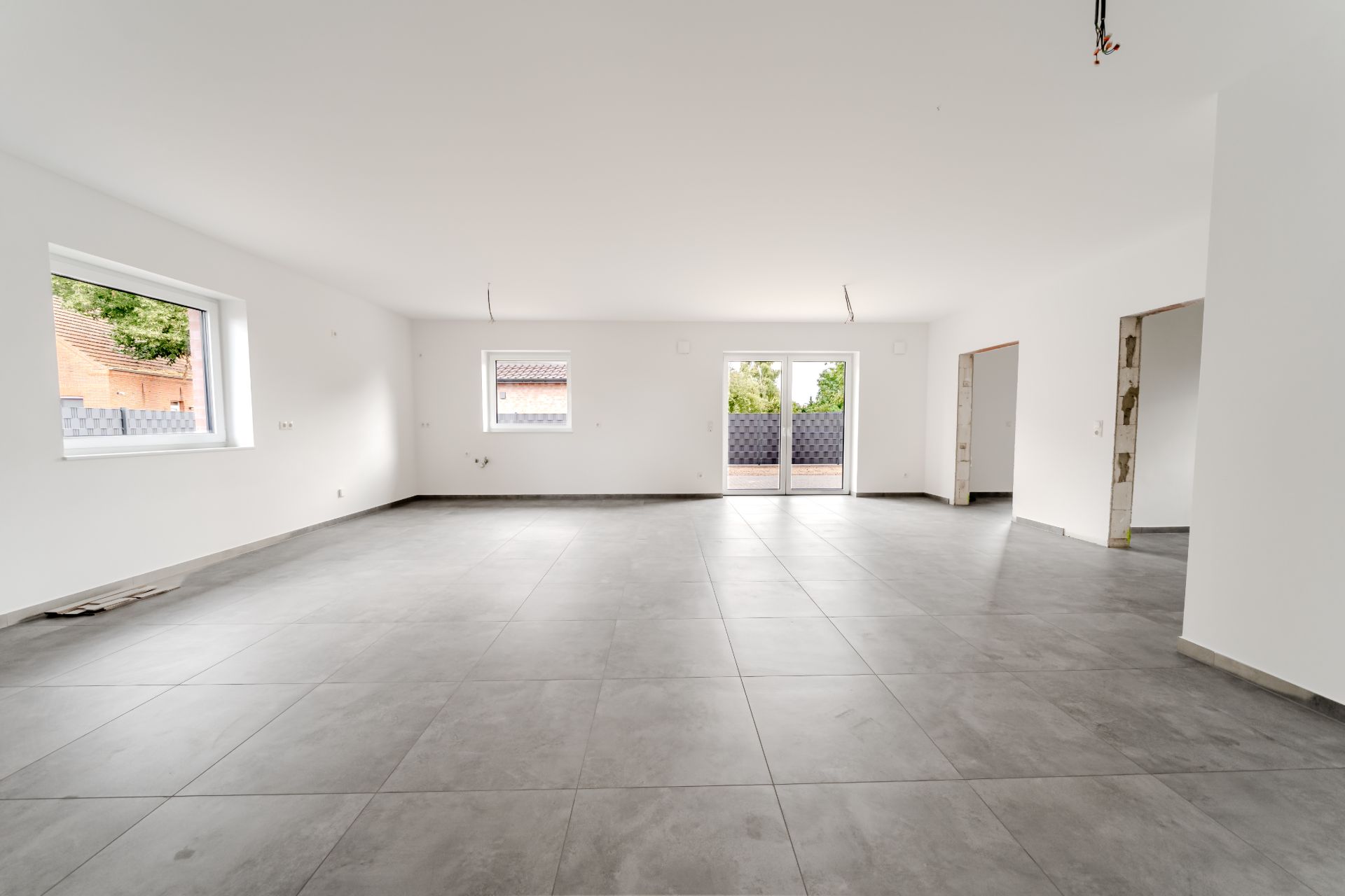 Großer, leerer Raum mit grauem Fliesenboden, weißen Wänden und Fenstern zur Terrasse.