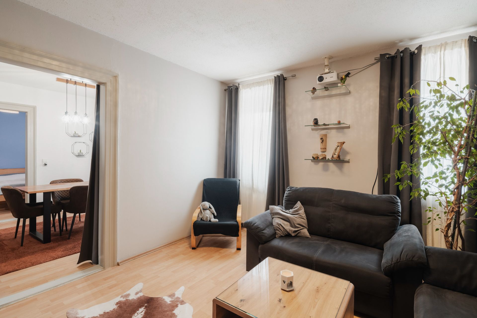 Wohnzimmer mit schwarzem Sofa, Sessel, Couchtisch, Pflanze und zwei Fenstern mit Vorhängen.