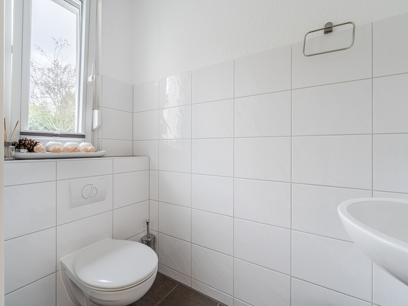Helles Badezimmer mit Wandtoilette, Fenster und Waschbecken, weiße Fliesen an den Wänden.