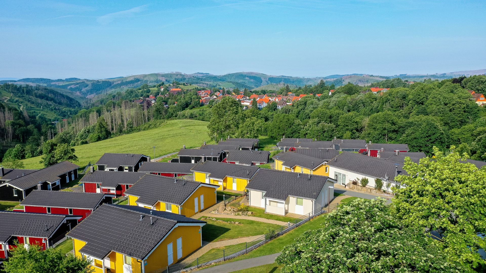 Mehrere bunte Einfamilienhäuser stehen auf einem grünen Hügel, im Hintergrund Wälder und Berge.
