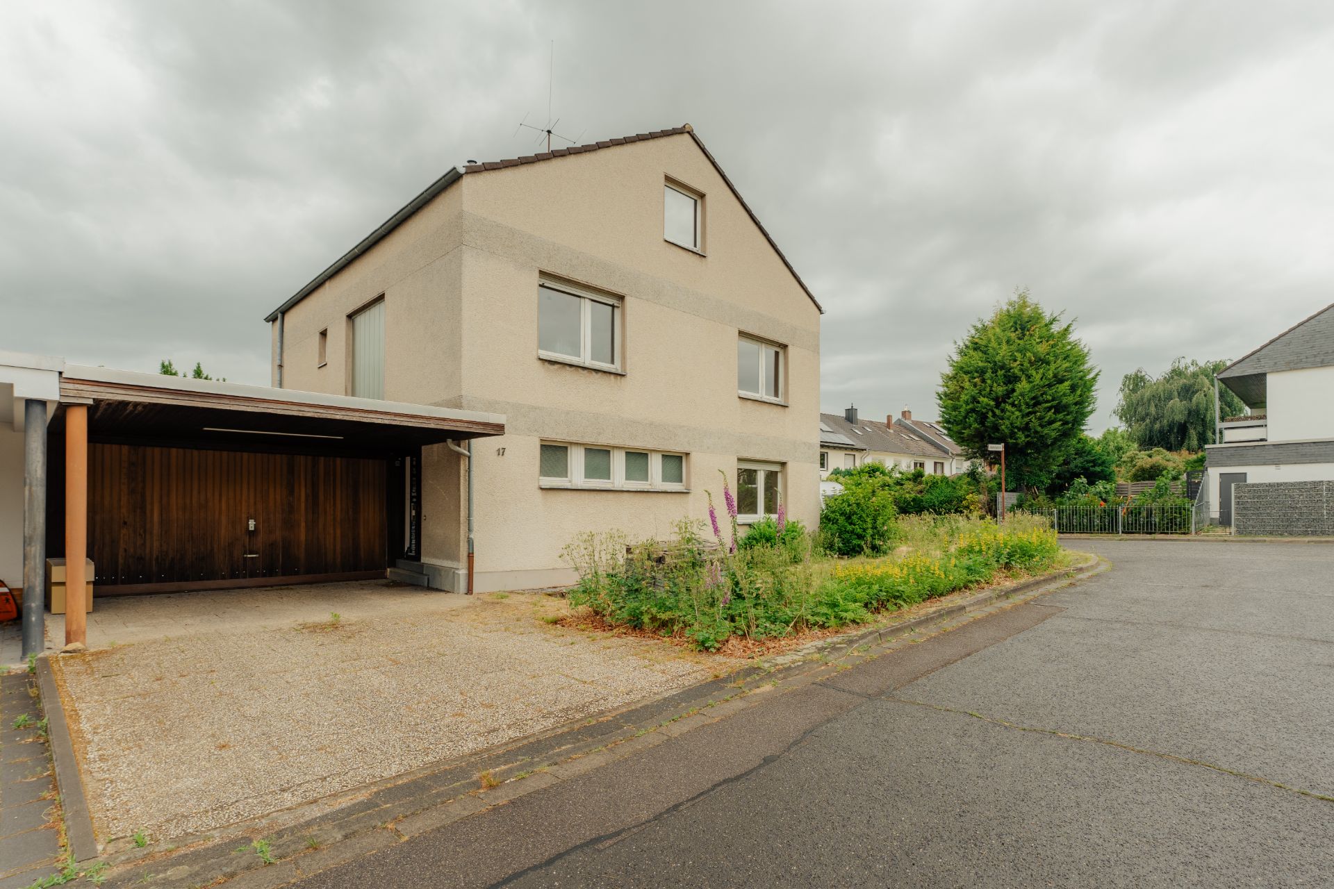 Zweistöckiges Haus mit Carport und Vorgarten an einer ruhigen Straße bei bewölktem Himmel.