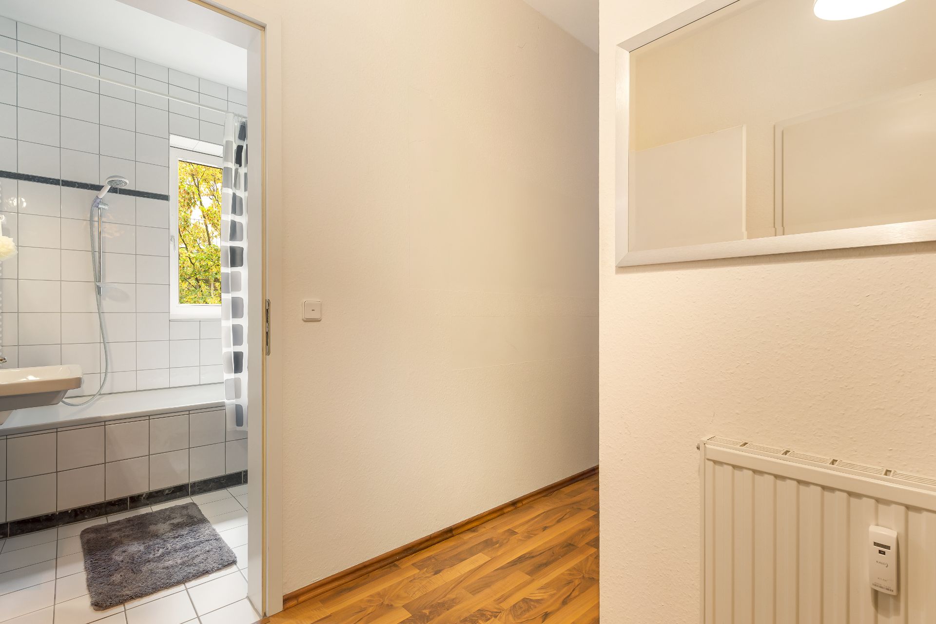 Heller Flur mit Parkettboden, daneben ein Badezimmer mit Fliesenwand und Fenster.