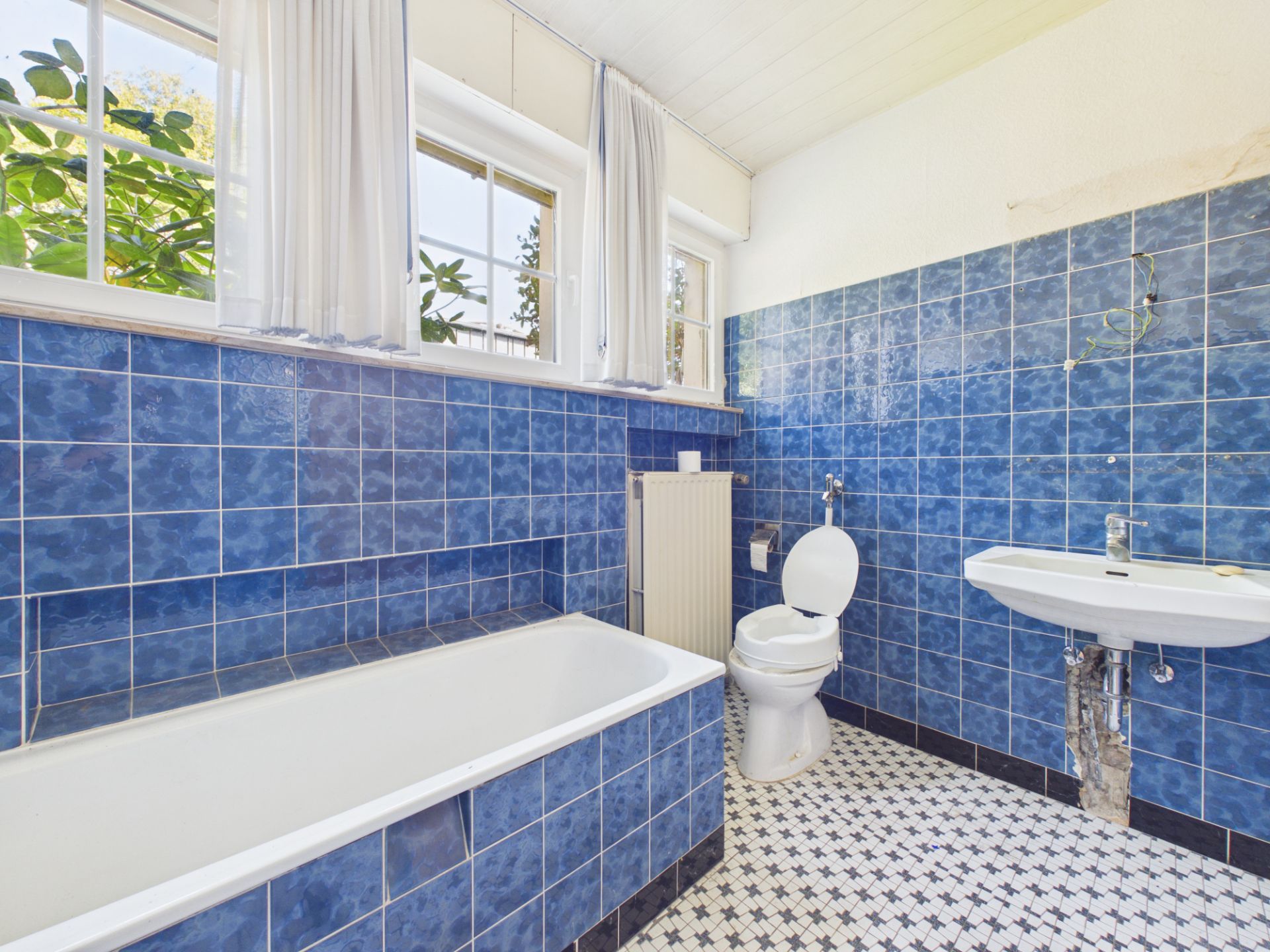 Badezimmer mit blauen Fliesen, Badewanne, Toilette, Waschbecken und Fenster mit Vorhängen.