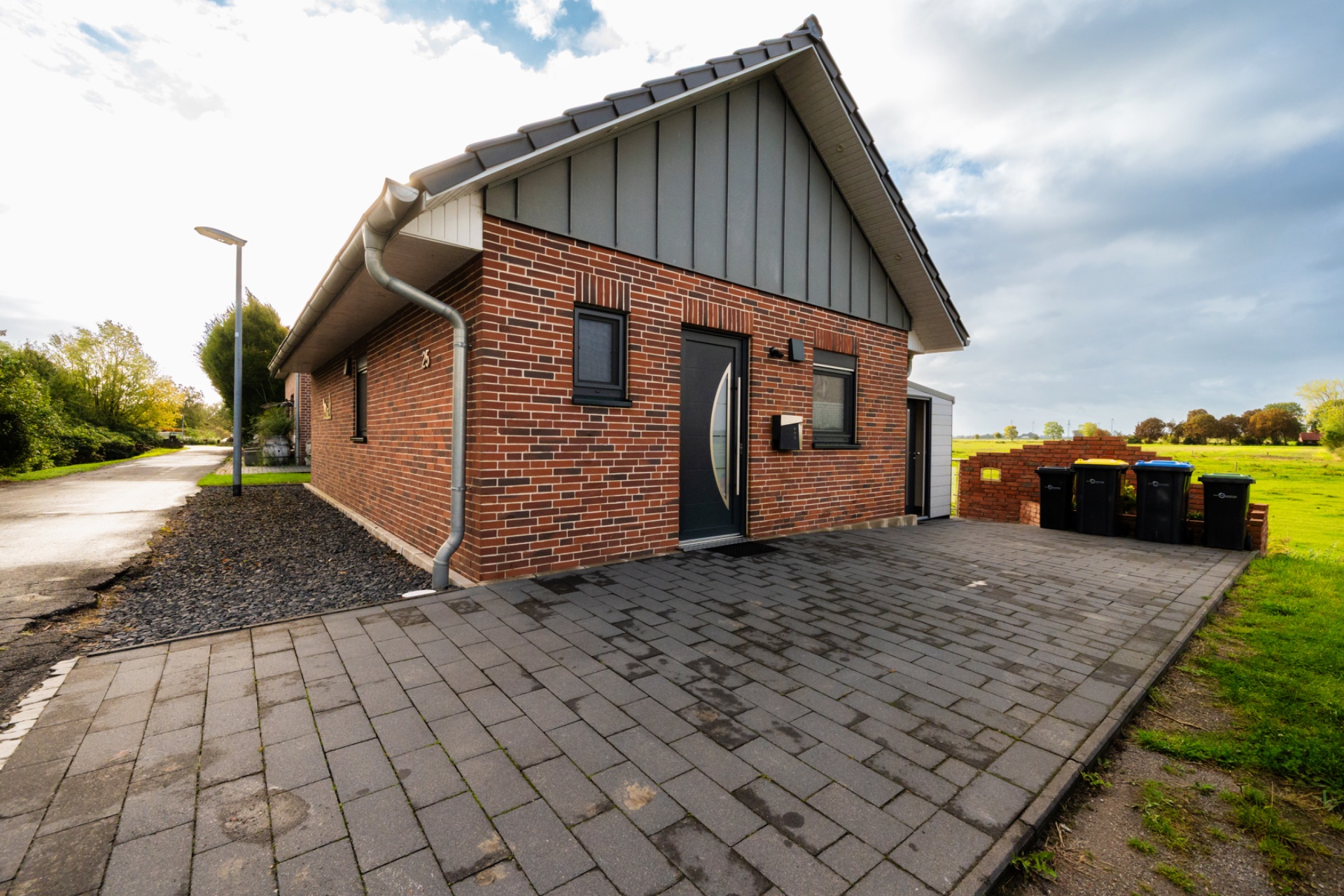 Backstein-Einfamilienhaus mit modernem Eingang und gepflasterter Einfahrt, mehrere Mülltonnen am Rand.