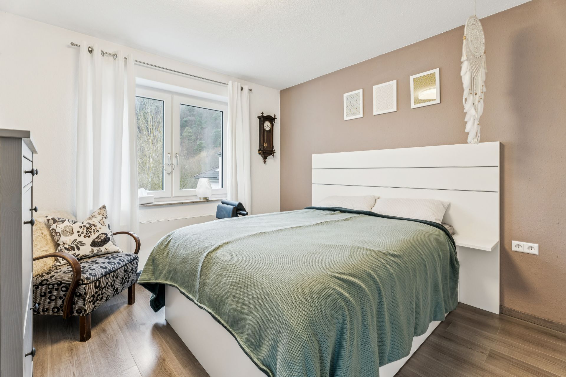 Helles Schlafzimmer mit großem Bett, Sessel, Fenster und Wanddeko.