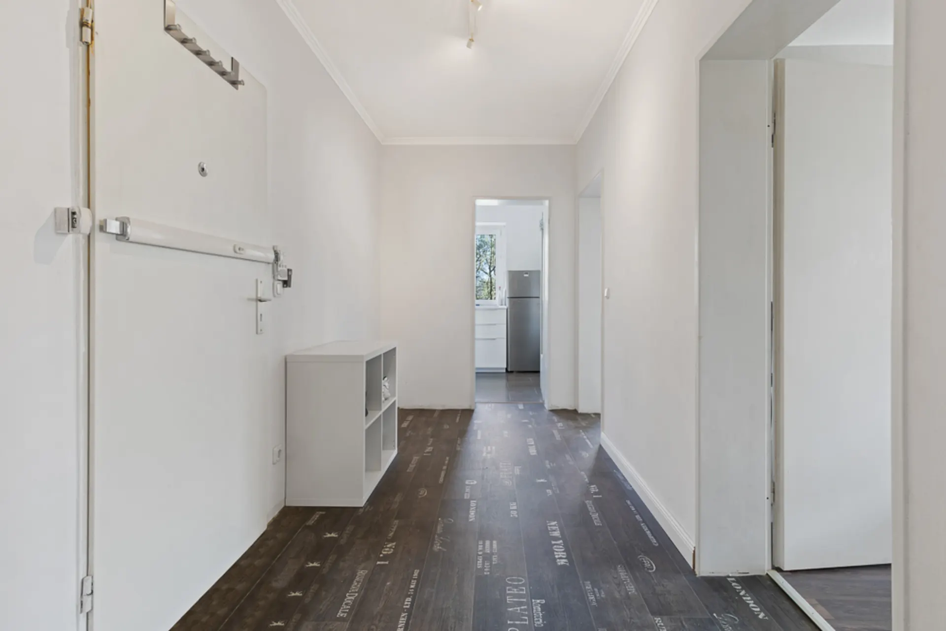 Heller Flur mit dunklem Boden, offener Tür zur Küche und Regal an der Wand.