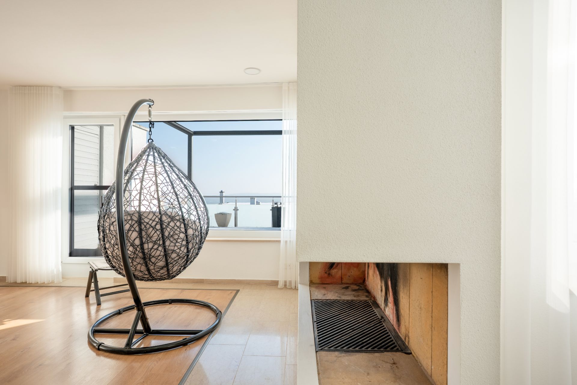 Hängesessel vor großem Fenster in modernem Wohnzimmer mit hellem Holzboden und Kamin.