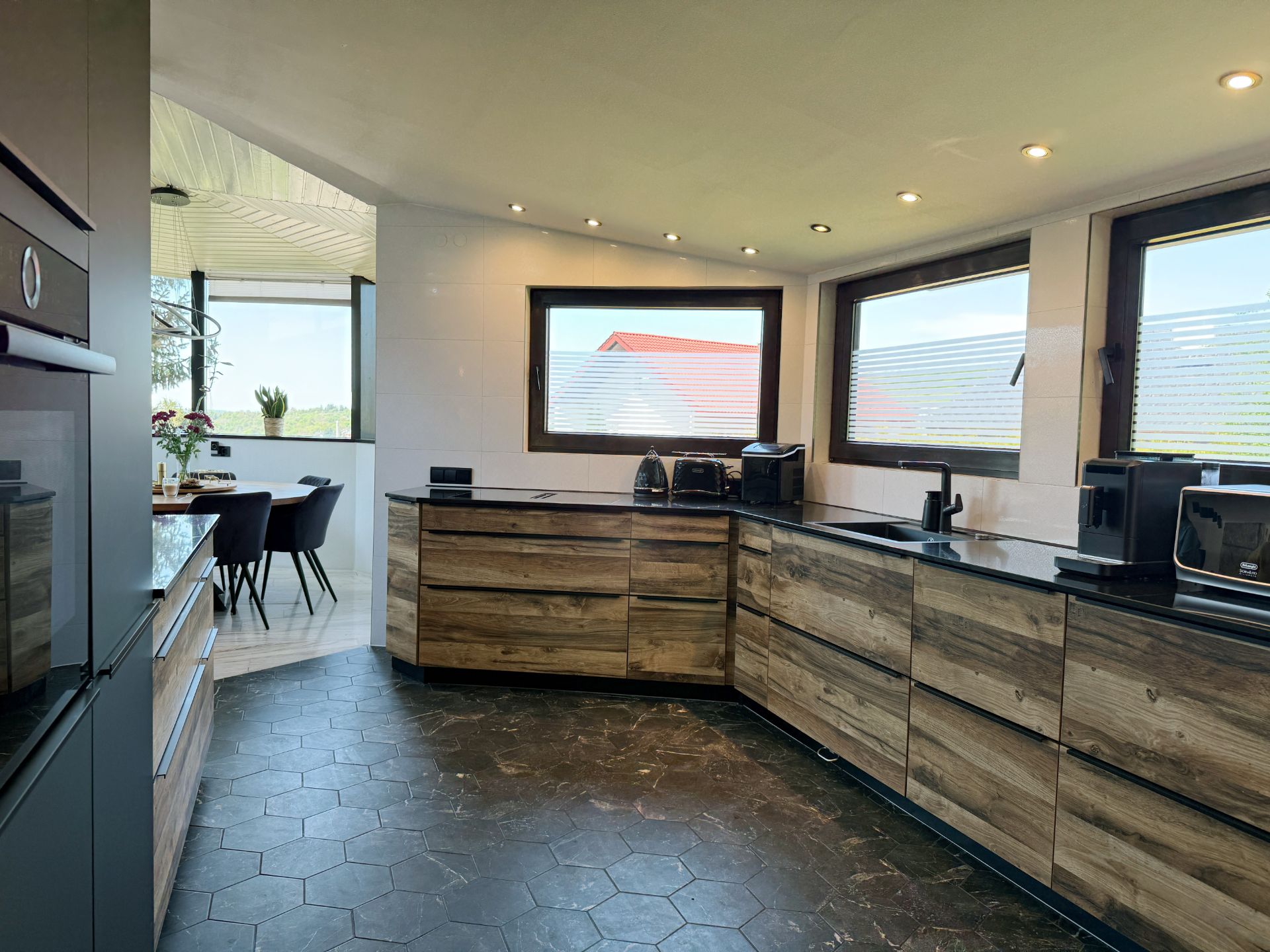 Moderne Küche mit Holzfronten, dunkler Arbeitsplatte und großen Fenstern, daneben ein Essbereich.