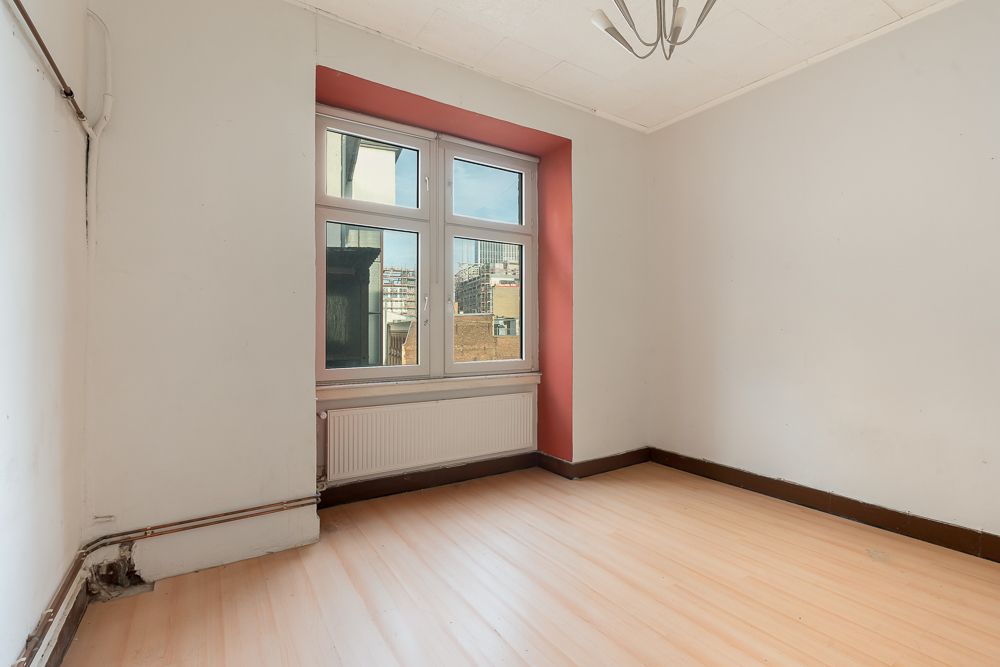 Leeres Zimmer mit großem Fenster, hellem Boden und weiß-grauen Wänden.