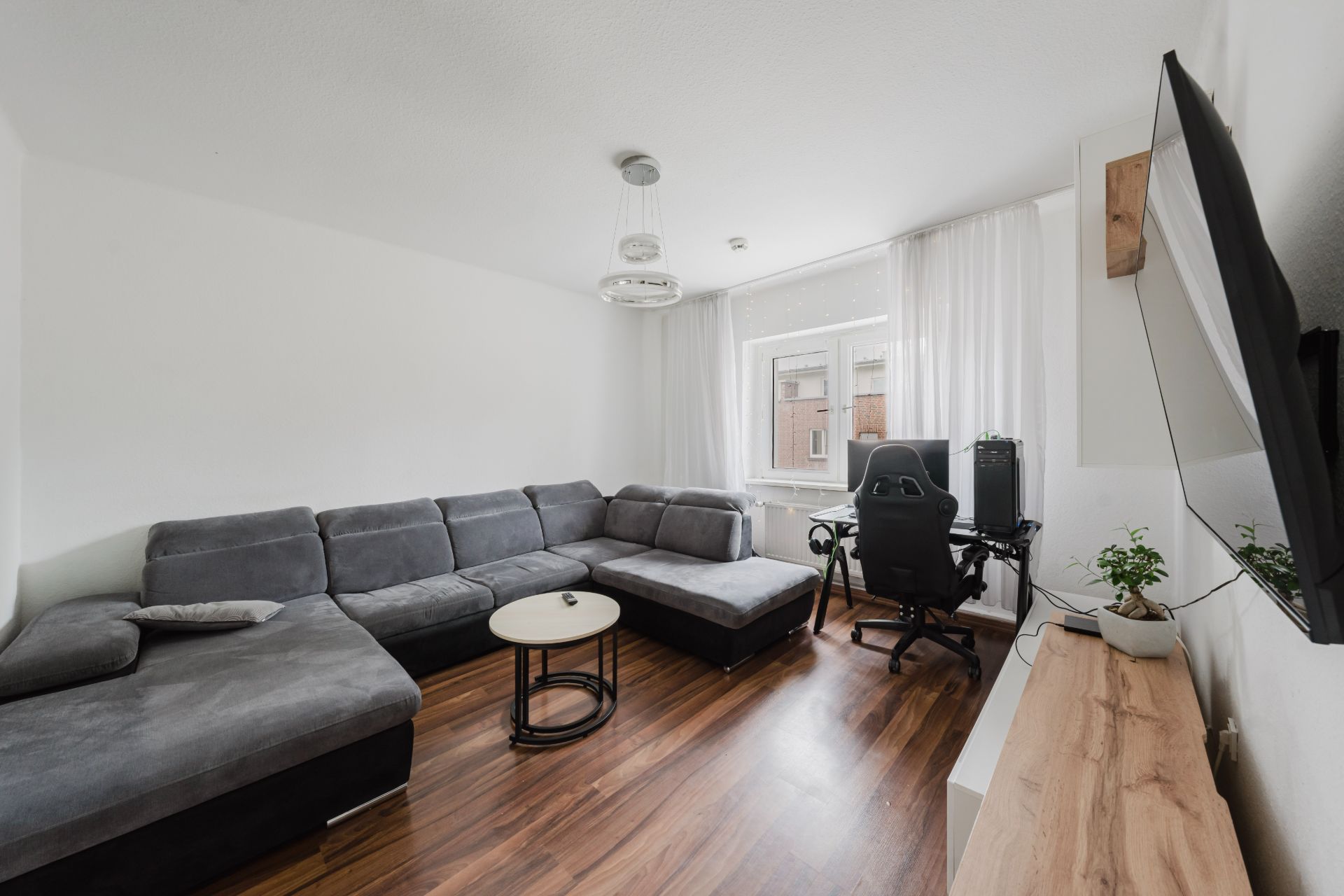Wohnzimmer mit grauem Ecksofa, Schreibtisch mit Stuhl, Fernseher und Holzboden.
