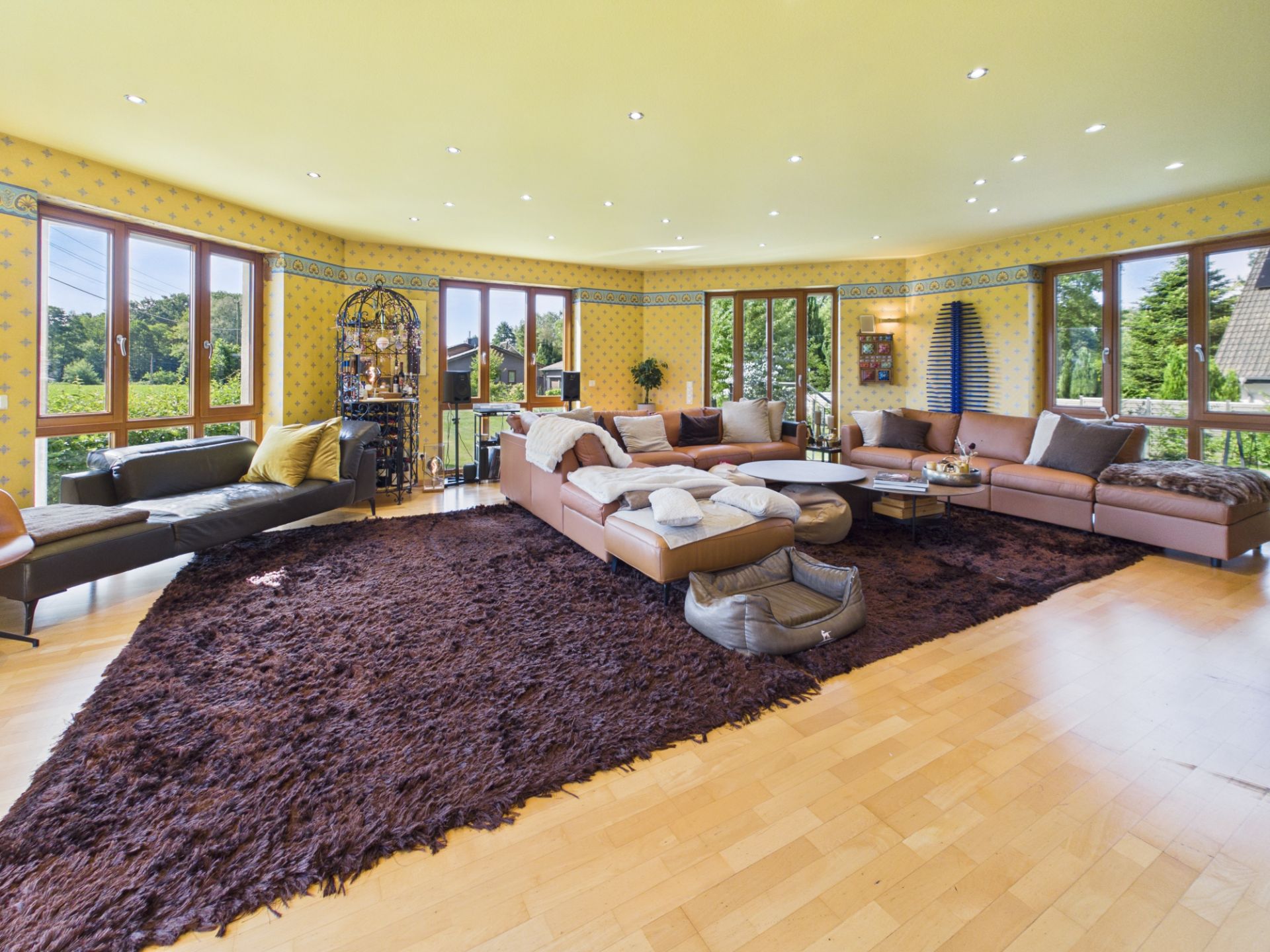 Großes Wohnzimmer mit braunem Teppich, Ecksofa und vielen Fenstern mit Blick ins Grüne.