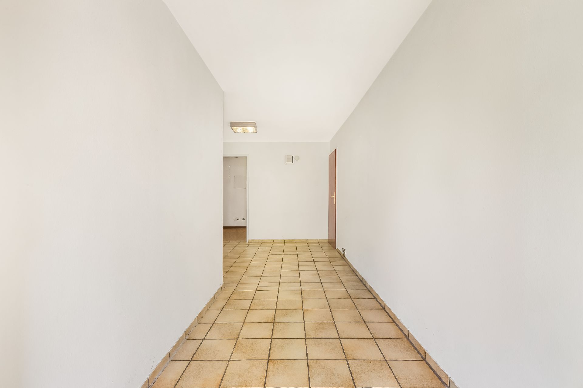 Flur mit beigem Fliesenboden und weißen Wänden, Blick auf eine Tür und einen offenen Durchgang.