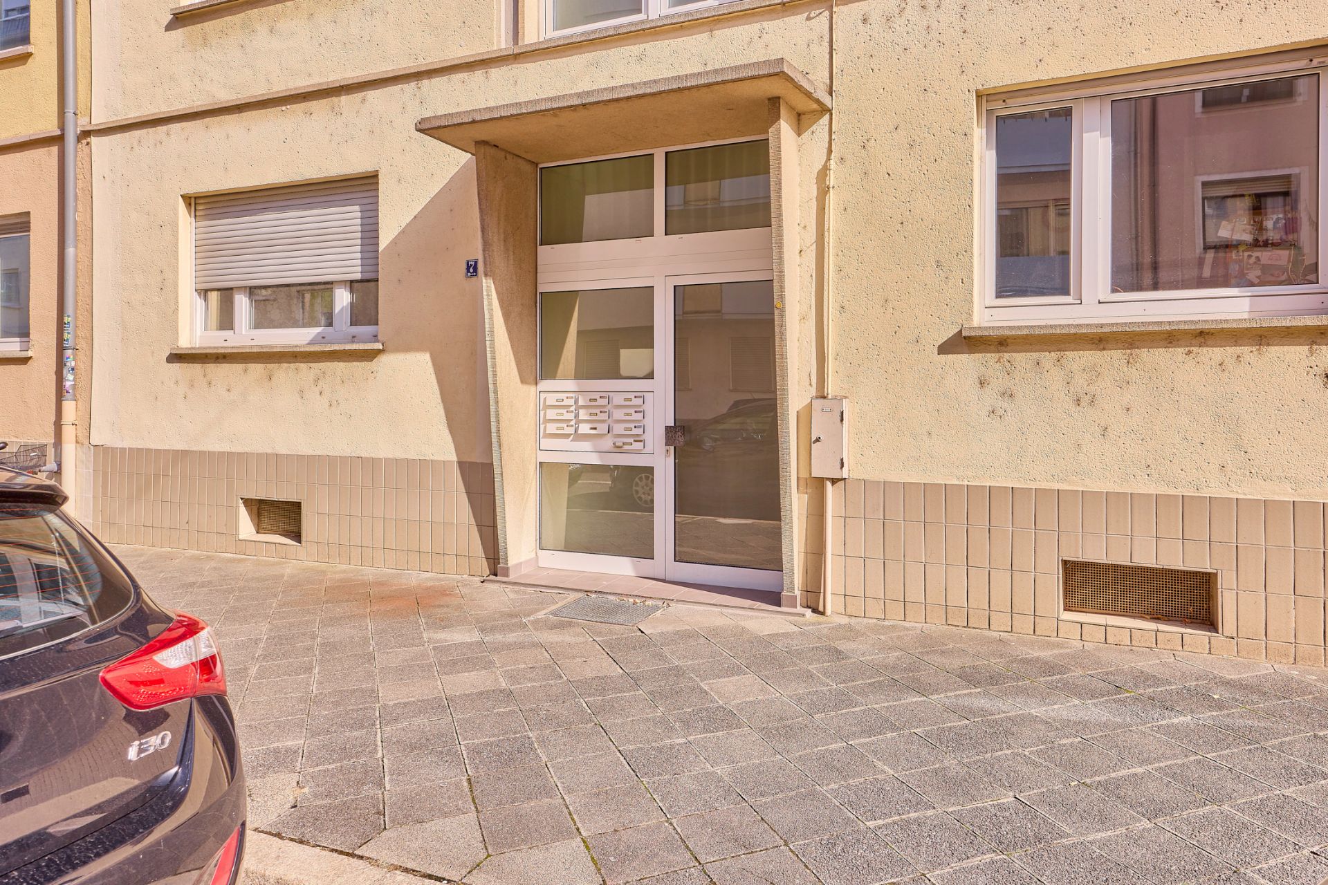 Eingang eines Mehrfamilienhauses mit Briefkästen neben der Tür, geparktes Auto links.