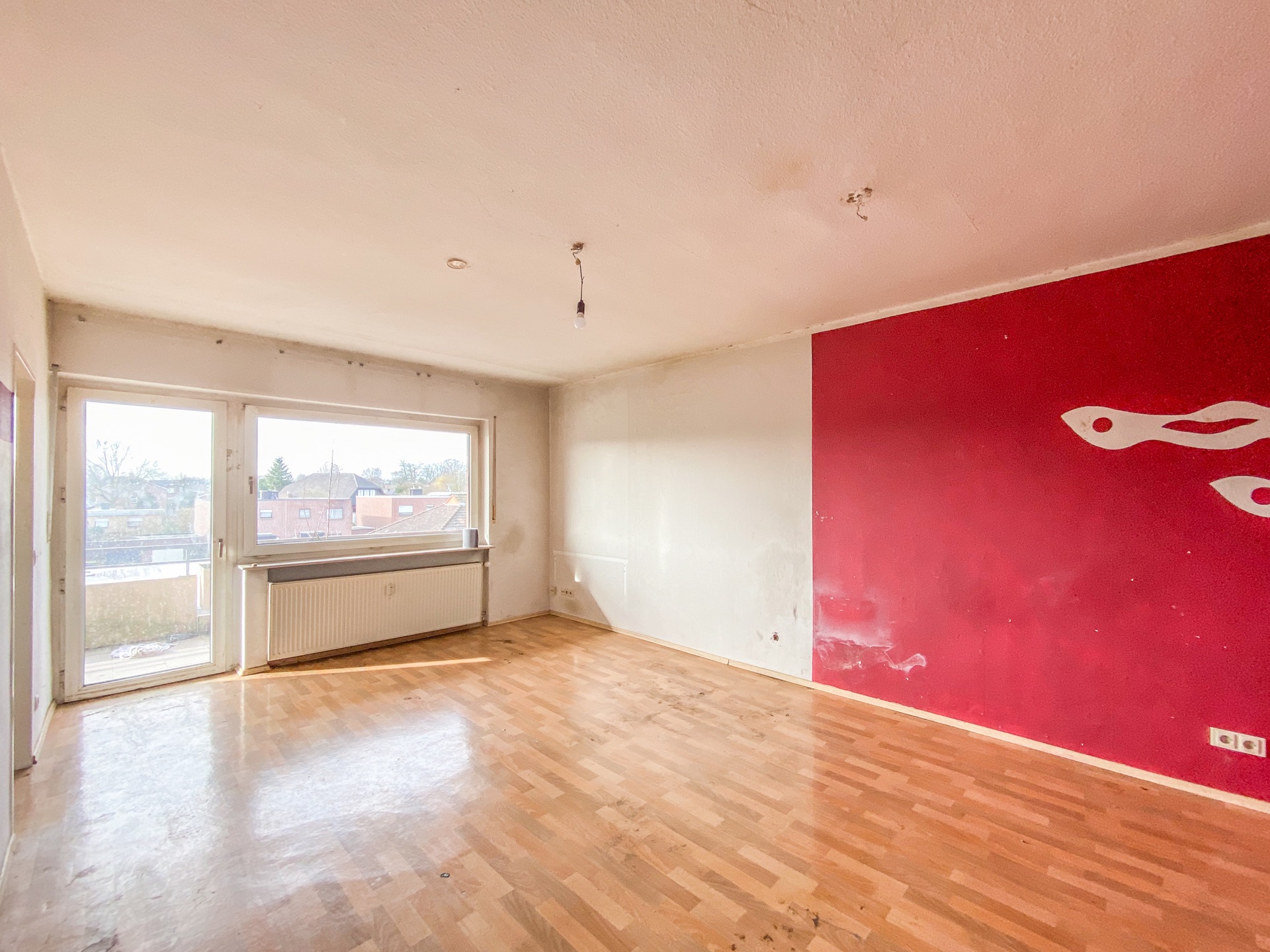 Leeres Zimmer mit großem Fenster, Parkettboden und roter Wand mit weißem Muster.