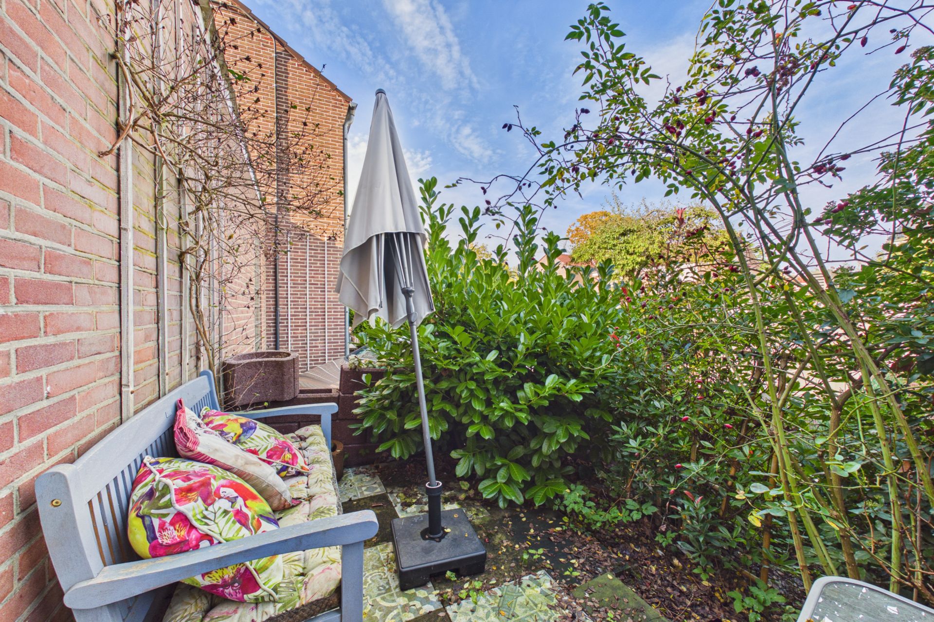 Kleine Terrasse mit Bank, bunten Kissen, geschlossenem Sonnenschirm und umgeben von grünen Pflanzen.