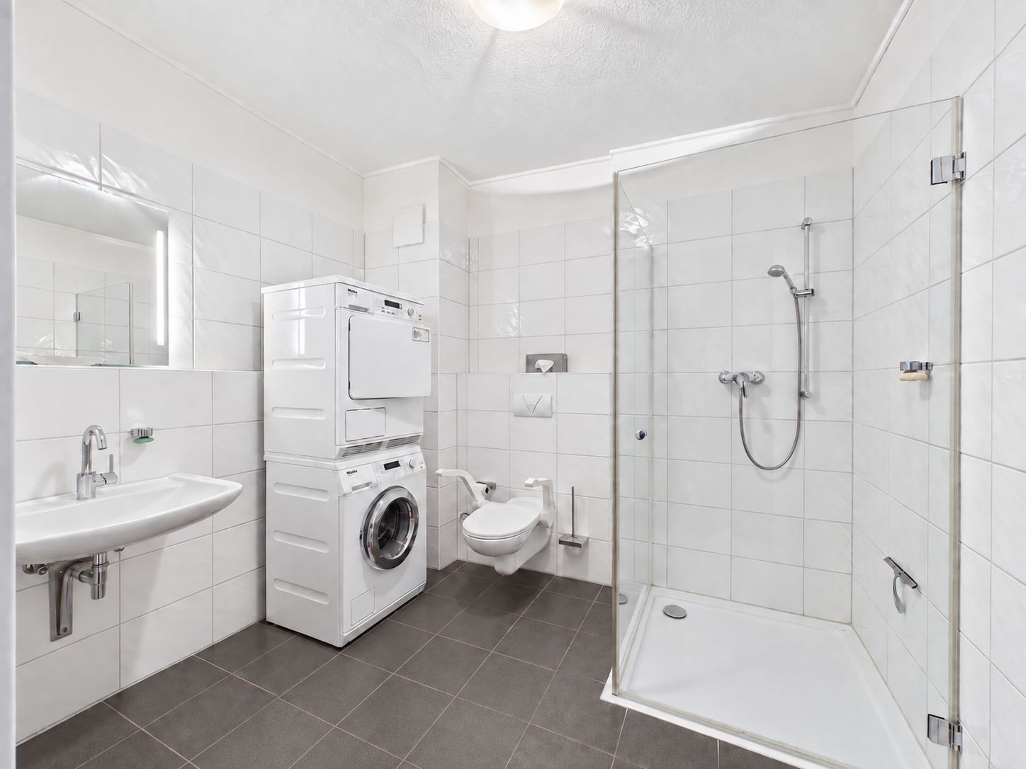 Helles Bad mit Dusche, Waschbecken, WC und gestapelter Waschmaschine und Trockner.