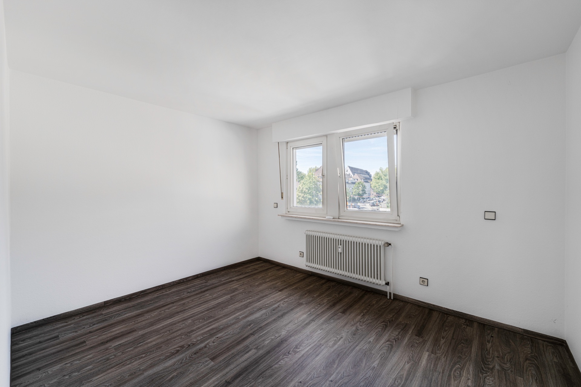 Leeres Zimmer mit weiß gestrichenen Wänden, dunklem Boden und zwei Fenstern mit Blick nach draußen.
