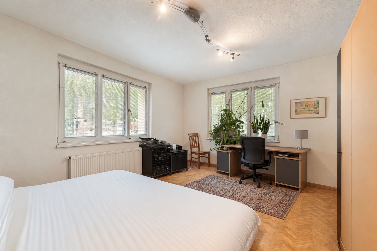 Helles Schlafzimmer mit Schreibtisch, Bett, Teppich und großen Fenstern, Zimmerpflanzen auf dem Schreibtisch.