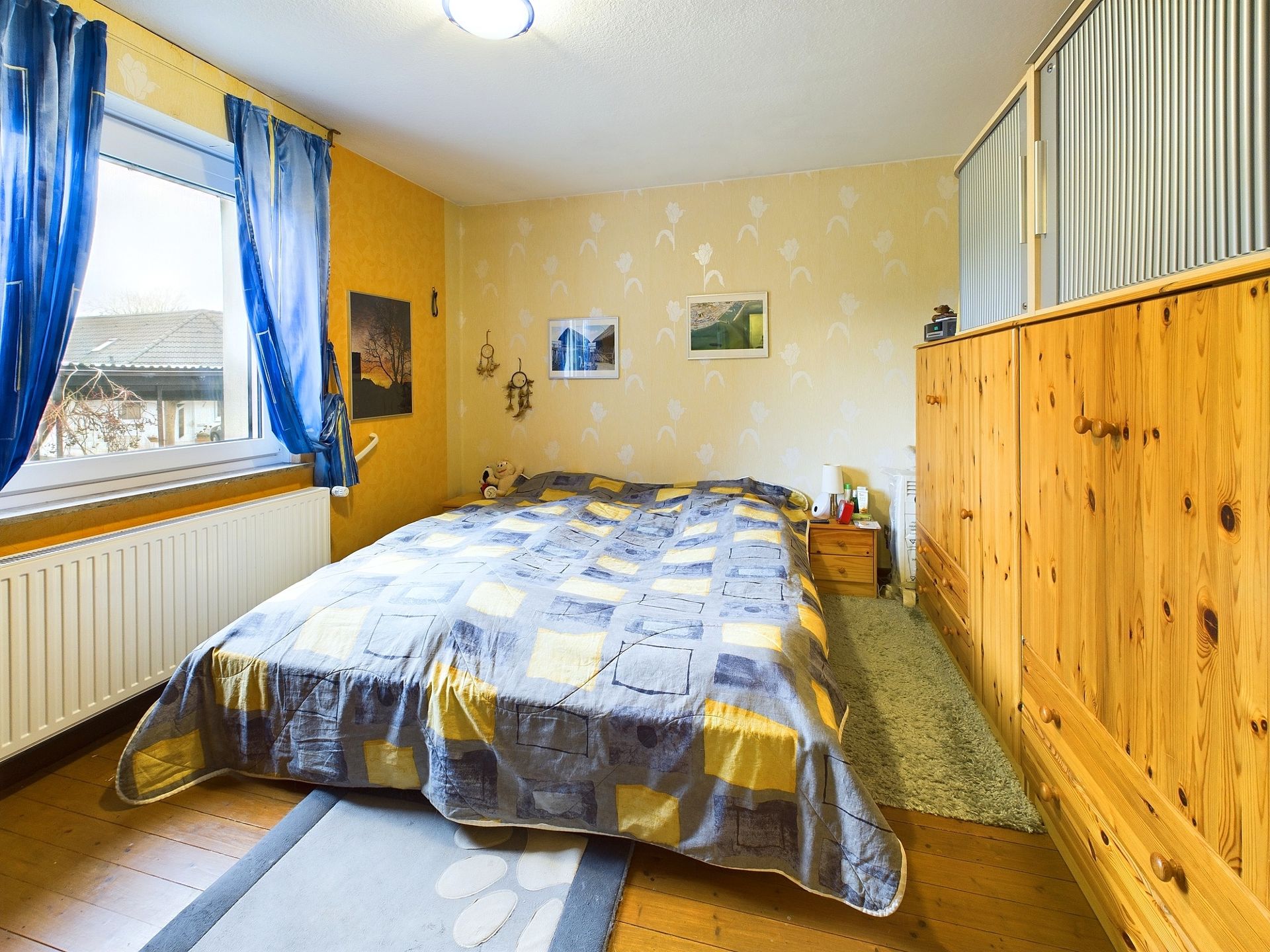 Schlafzimmer mit Doppelbett, Holzmöbeln, blau-gelben Vorhängen und Bildern an der Wand.