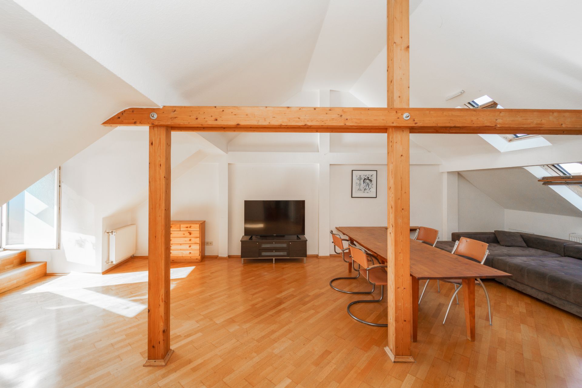 Helles Wohnzimmer mit Holzbalken, Esstisch, Stühlen, Sofa und Fernseher unter schräger Decke.