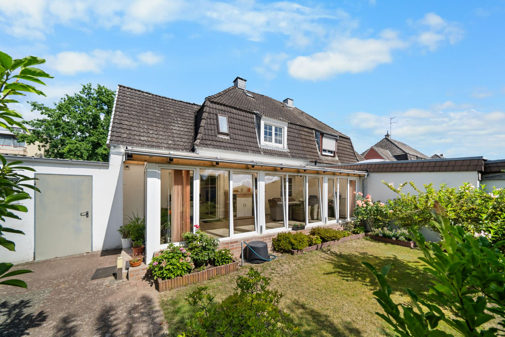 Haus mit verglaster Terrasse, Garten und Blumenbeeten bei sonnigem Wetter.