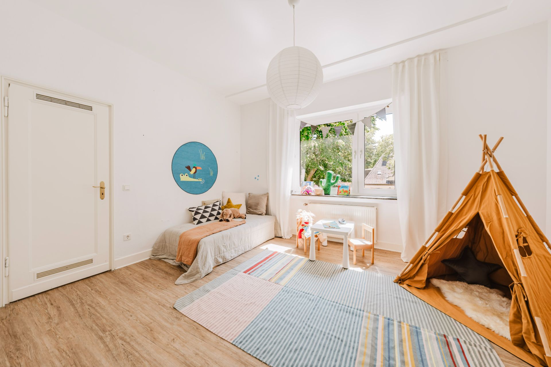 Helles Kinderzimmer mit Teppich, Bett, Spielzelt und kleinem Tisch vor einem Fenster.