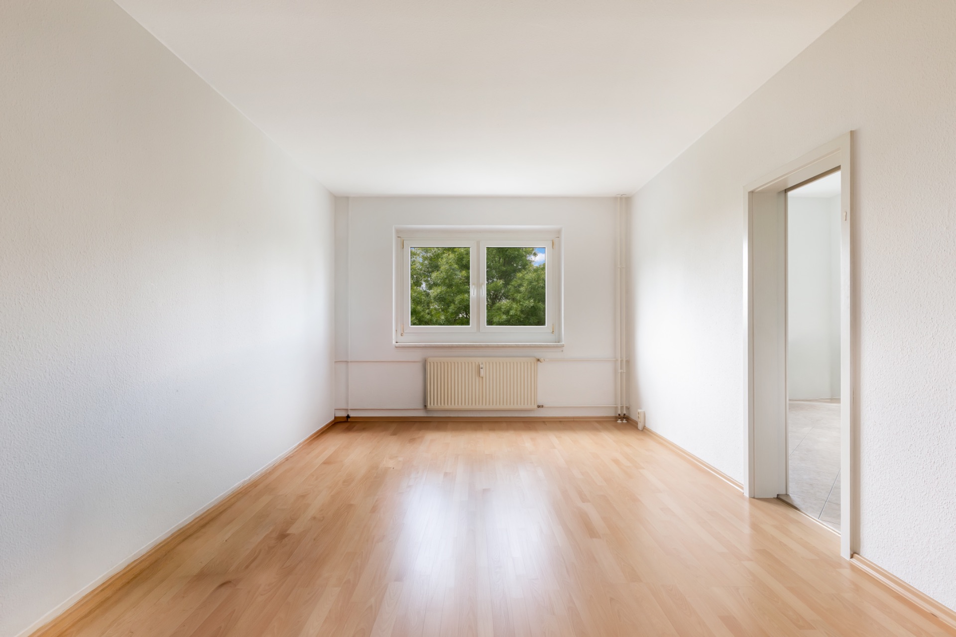 Leeres Zimmer mit hellem Holzfußboden, weißen Wänden und Fenster mit Blick ins Grüne.