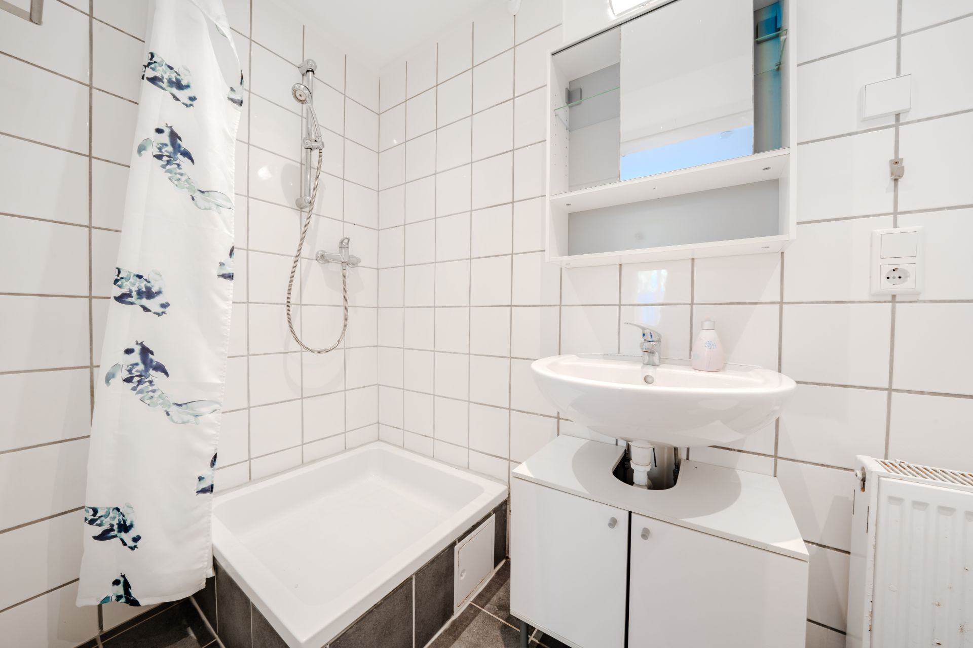Helles Bad mit Dusche, Duschvorhang, Waschbecken, Spiegelschrank und weißen Fliesen.