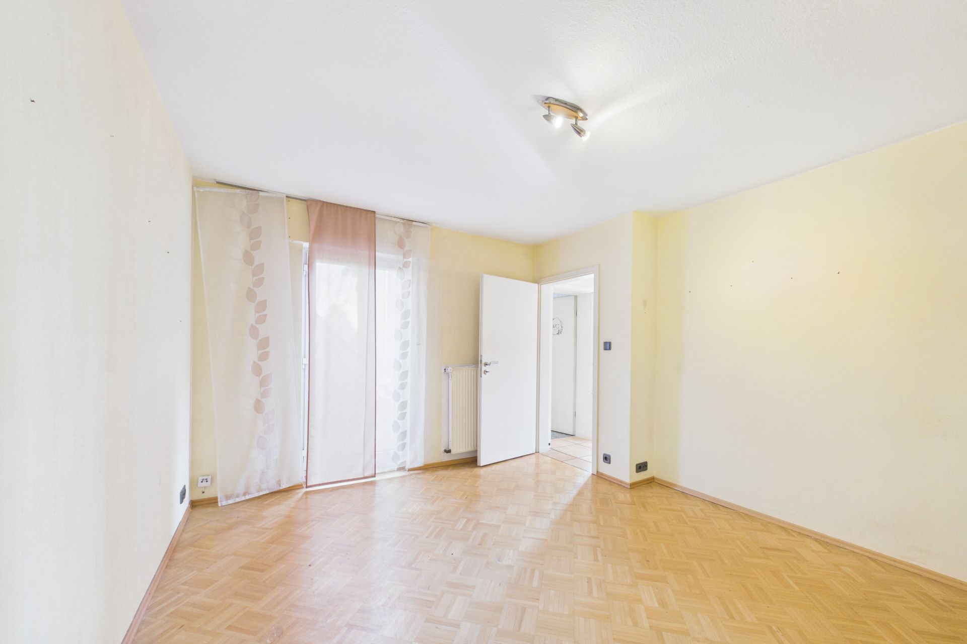 Heller leerer Raum mit Parkettboden, großem Fenster mit Vorhängen und offener Tür.