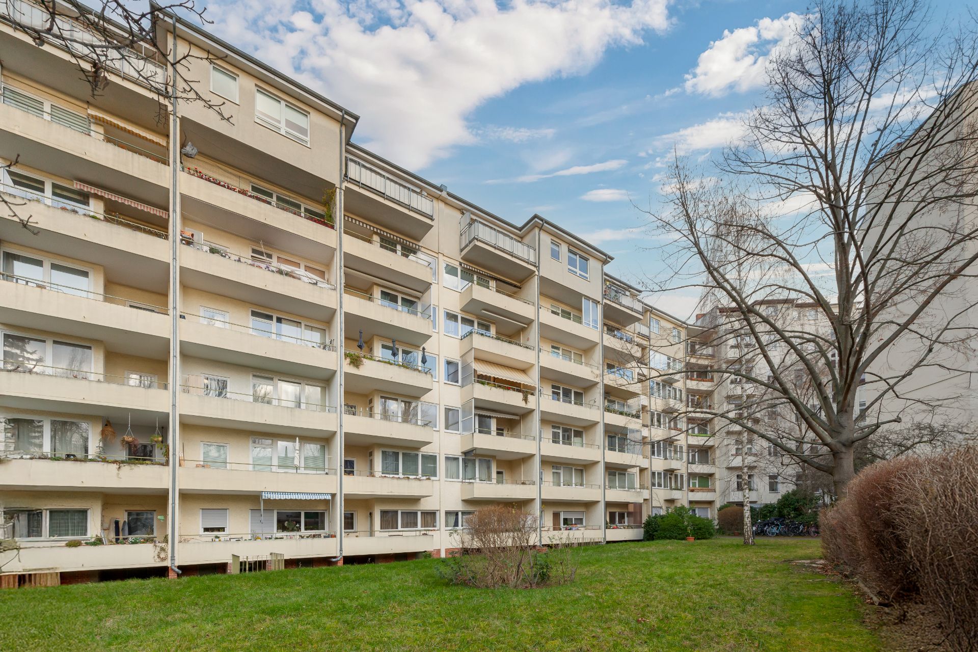 Mehrstöckiges Wohnhaus mit Balkonen und Vorgarten, großer Baum rechts im Bild.