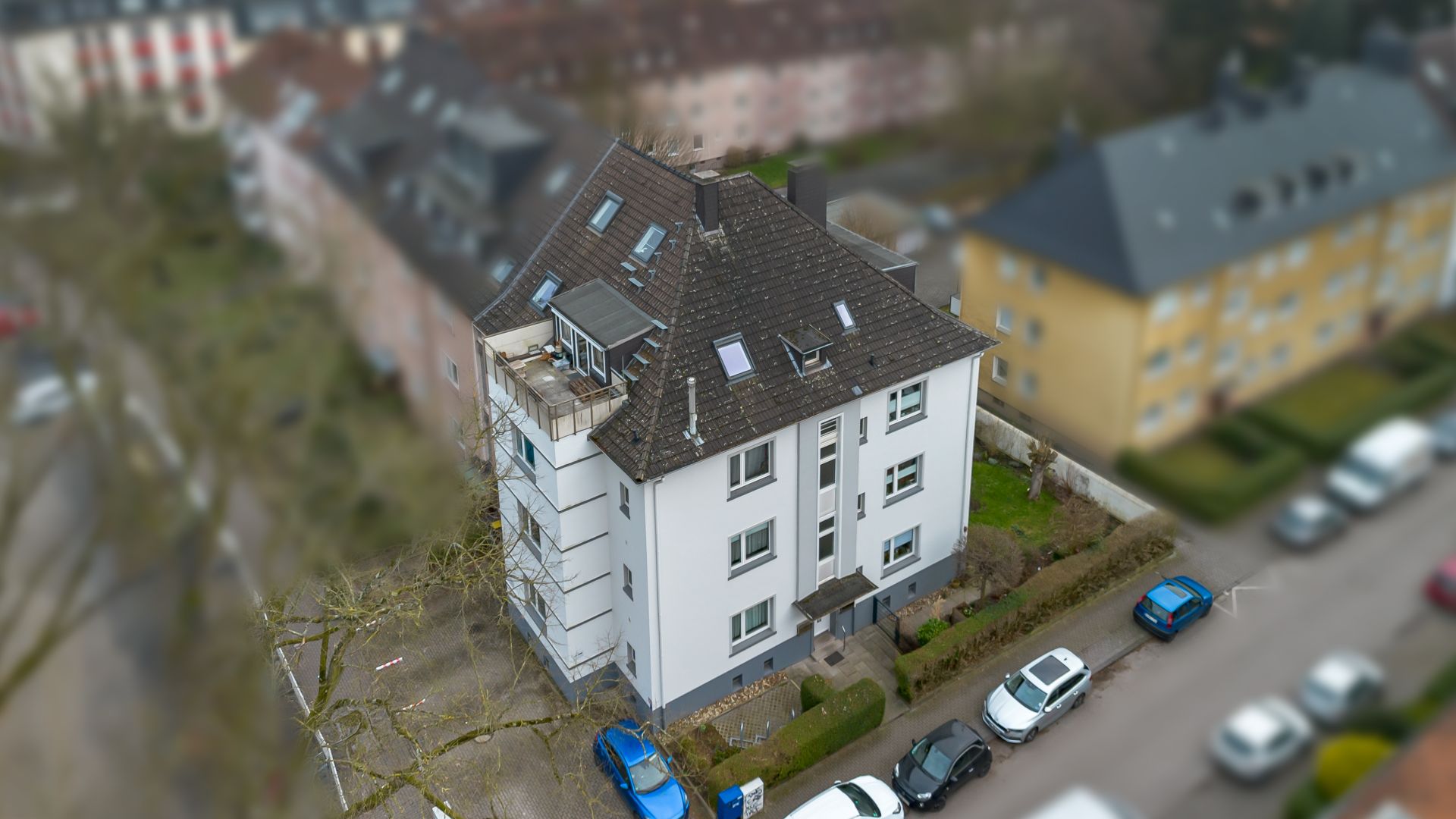 Mehrstöckiges Wohnhaus mit Balkon, Parkplatz und angrenzendem Gehweg von oben gesehen.