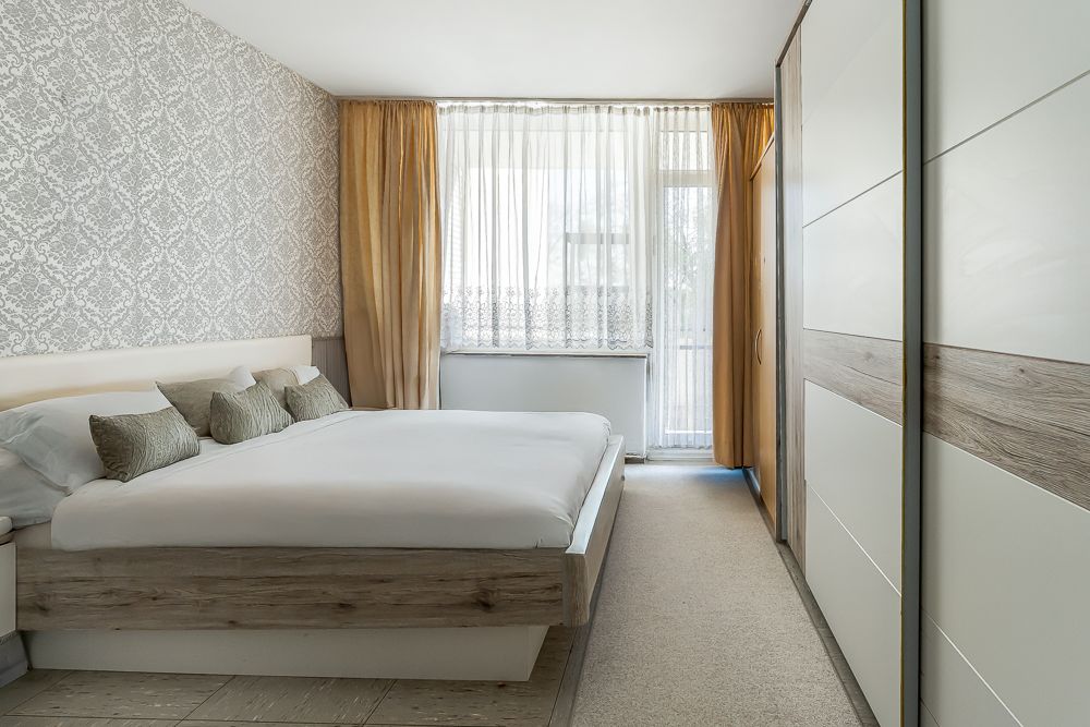 Helles Schlafzimmer mit Doppelbett, Kleiderschrank und großen Fenstern mit Vorhängen.