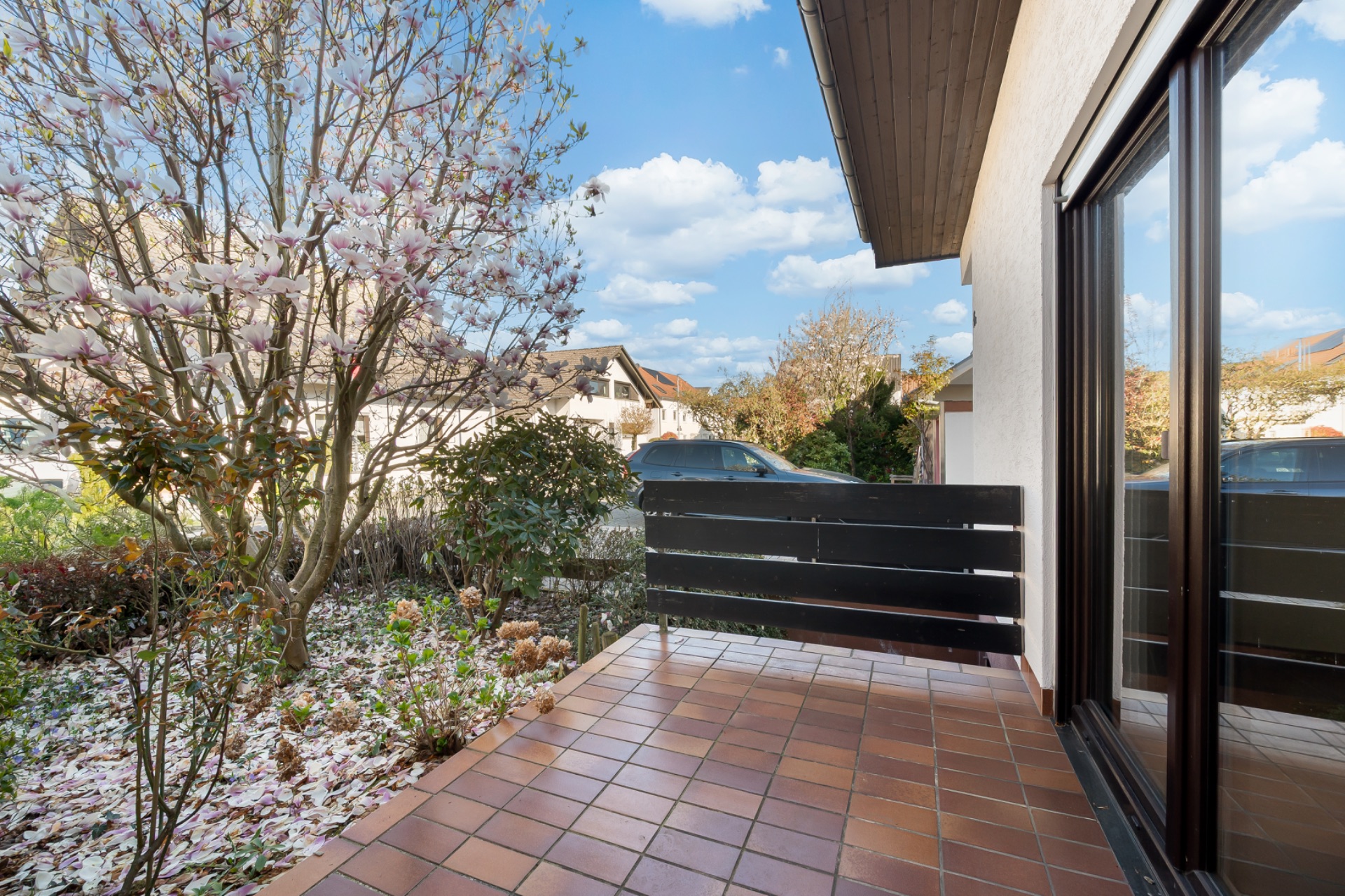 Geflieste Terrasse mit Blumenbeet, Baum und schwarzem Geländer an einem Wohnhaus.