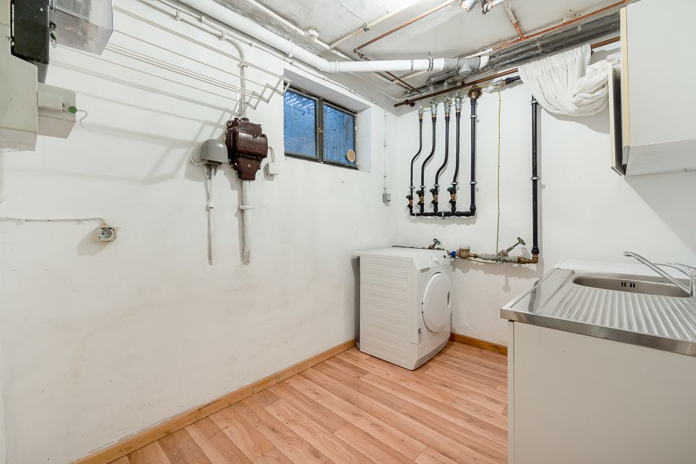 Waschraum mit Waschmaschine, Spülbecken, Rohren an der Wand und kleinem Fenster.